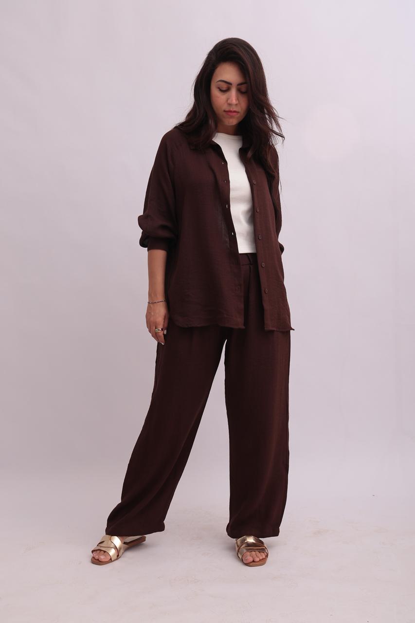 Full-body shot of a model wearing an open chocolate brown linen overshirt over a white tank top, paired with matching dark brown linen trousers.