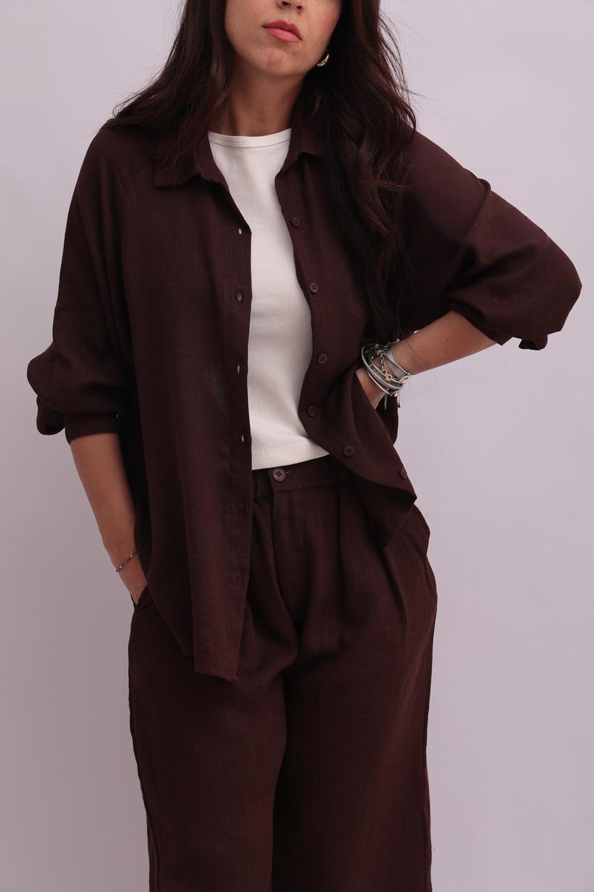 Close-up shot of a model wearing a chocolate brown linen overshirt styled open with a white tank top and matching linen trousers.