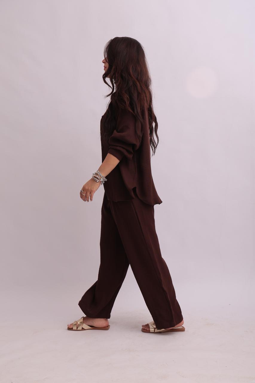 Side profile of a model walking while wearing a matching chocolate brown linen overshirt and wide-leg trousers.