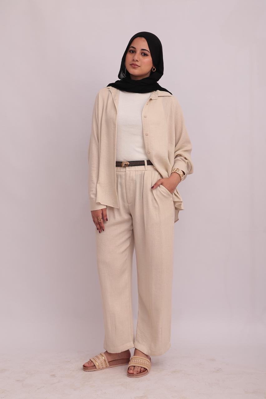 Full-body shot of a model in a black hijab, wearing an open cream linen overshirt over a white top, styled with cream wide-leg trousers.